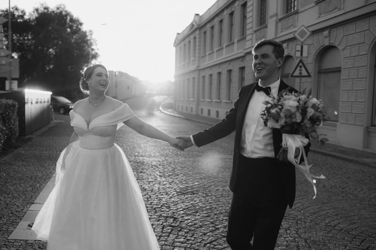 Czarno-białe zdjęcie uśmiechniętej pary młodej, idącej za rękę po brukowanej ulicy miejskiej w promieniach zachodzącego słońca, panna młoda w sukni z odkrytymi ramionami, pan młody w smokingu...