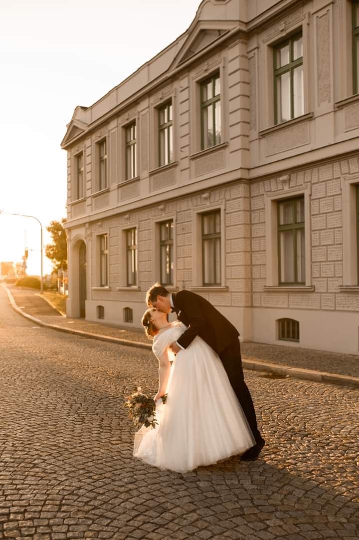 Para młoda całująca się na brukowanej ulicy w promieniach zachodzącego słońca, na tle budynku z klasyczną architekturą.