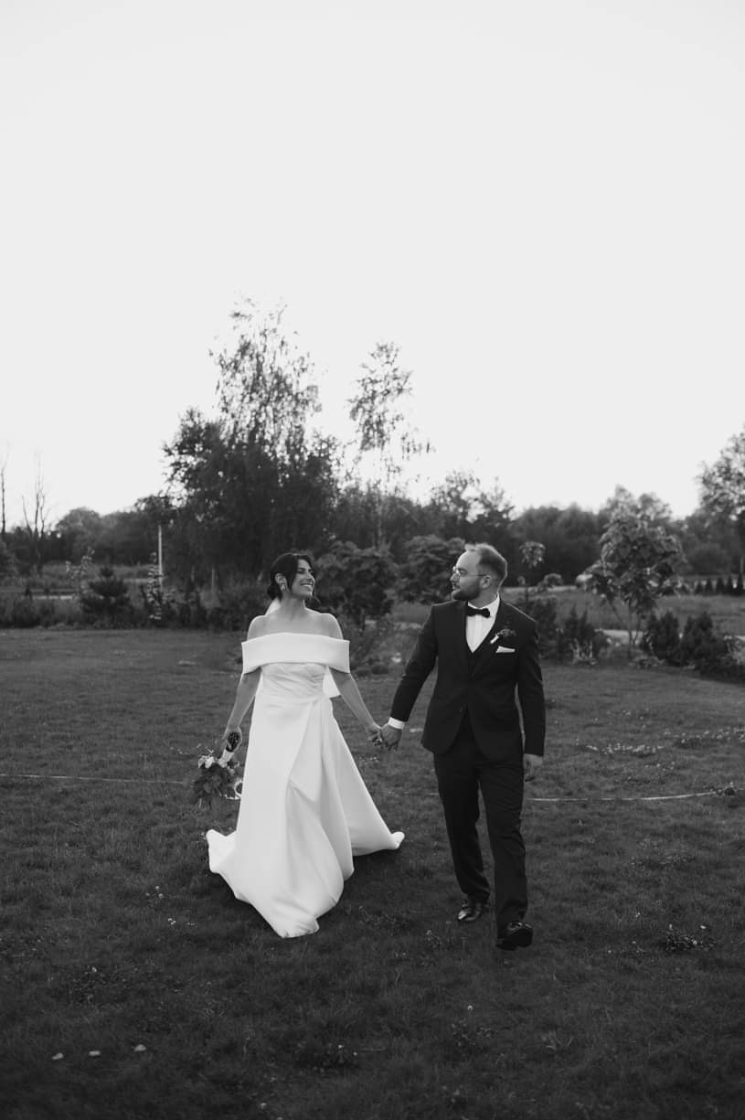 Czarno-białe zdjęcie pary młodej idącej po trawniku, panna młoda w sukni z odkrytymi ramionami trzyma bukiet, pan młody w garniturze z muszką, drzewa w tle.