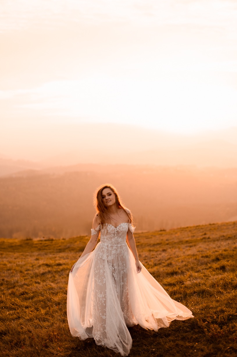 Uśmiechnięta kobieta w zwiewnej, koronkowej sukni spaceruje po wzgórzu o zachodzie słońca, z widokiem na zamglone góry w tle.