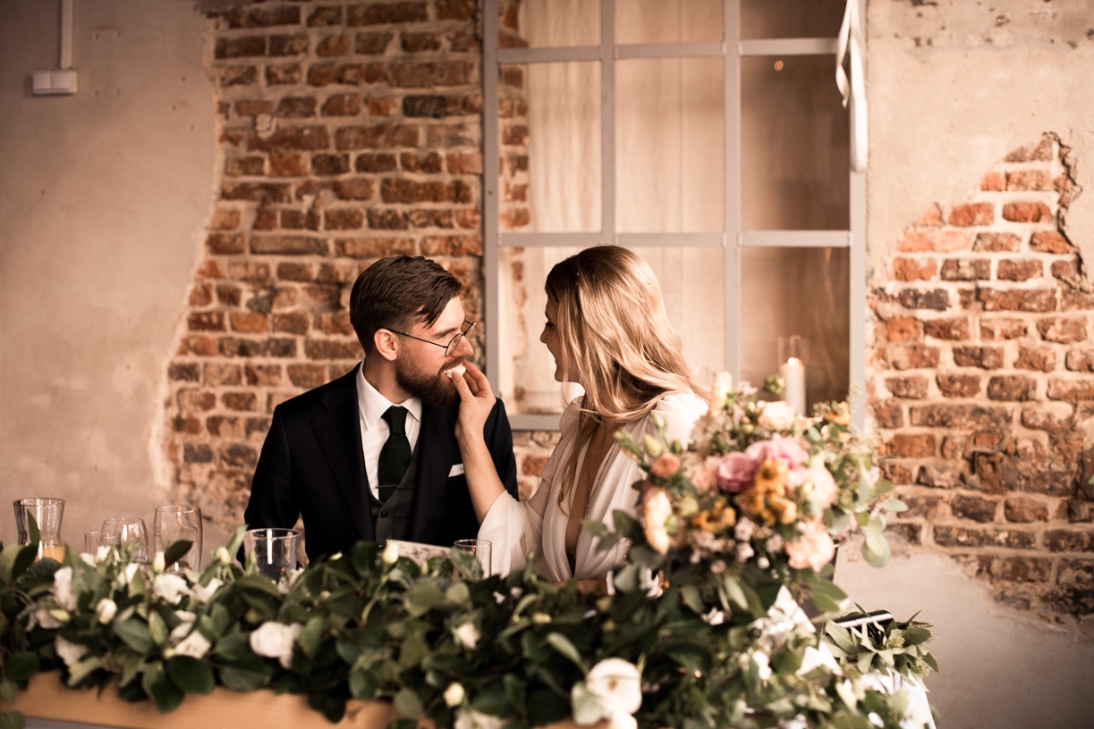 Panna młoda karmi pana młodego przy stole udekorowanym kwiatami na tle ceglanej ściany i okna podczas przyjęcia weselnego.
