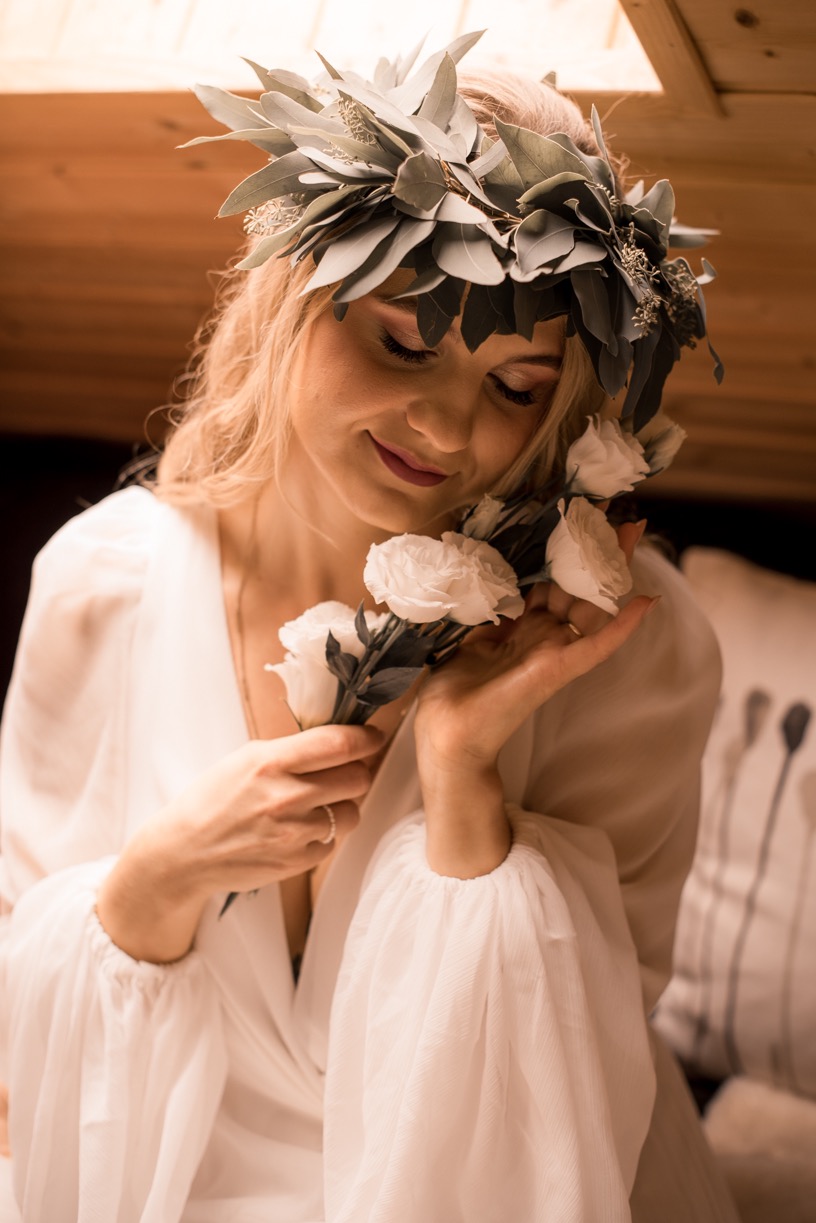 Portret kobiety w białej sukience i wianku z eukaliptusa, trzymającej białe róże, z delikatnym uśmiechem i zamkniętymi oczami, w ciepłym oświetleniu wnętrza.