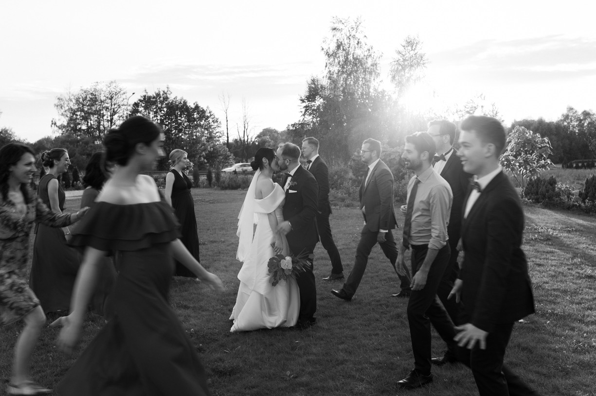 Czarno-białe zdjęcie całującej się pary młodej na tle zielonego trawnika, otoczonej przez gości weselnych w strojach wieczorowych, z delikatnym światłem słonecznym w tle.