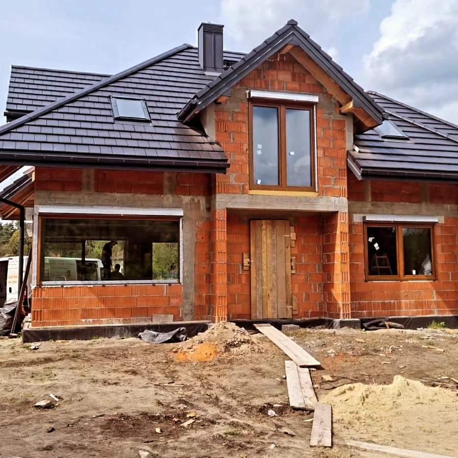 Dom w trakcie budowy z zamontowanymi oknami w brązowych ramach i szarymi roletami zewnętrznymi, widoczne cegły i drewniane deski przed wejściem.