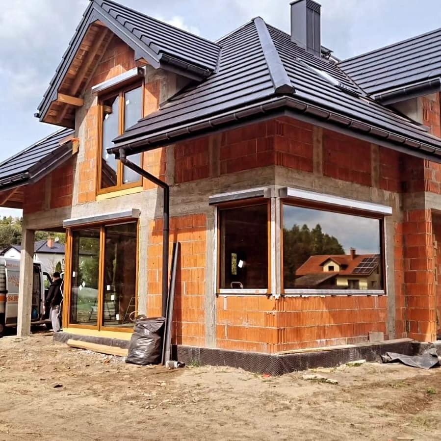 Nowy dom w trakcie budowy z zamontowanymi oknami z brązowymi ramami, widoczne rolety zewnętrzne i czarny worek gruzu przy ścianie.