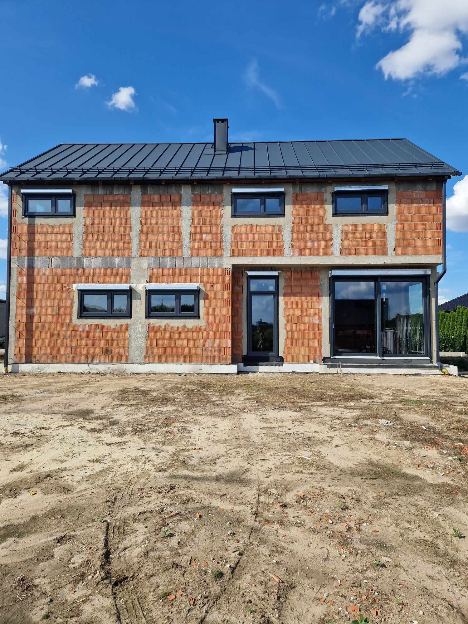 Surowy stan budynku z czarnymi oknami i drzwiami tarasowymi, widoczne szare rolety zewnętrzne i ceglana elewacja w trakcie budowy.