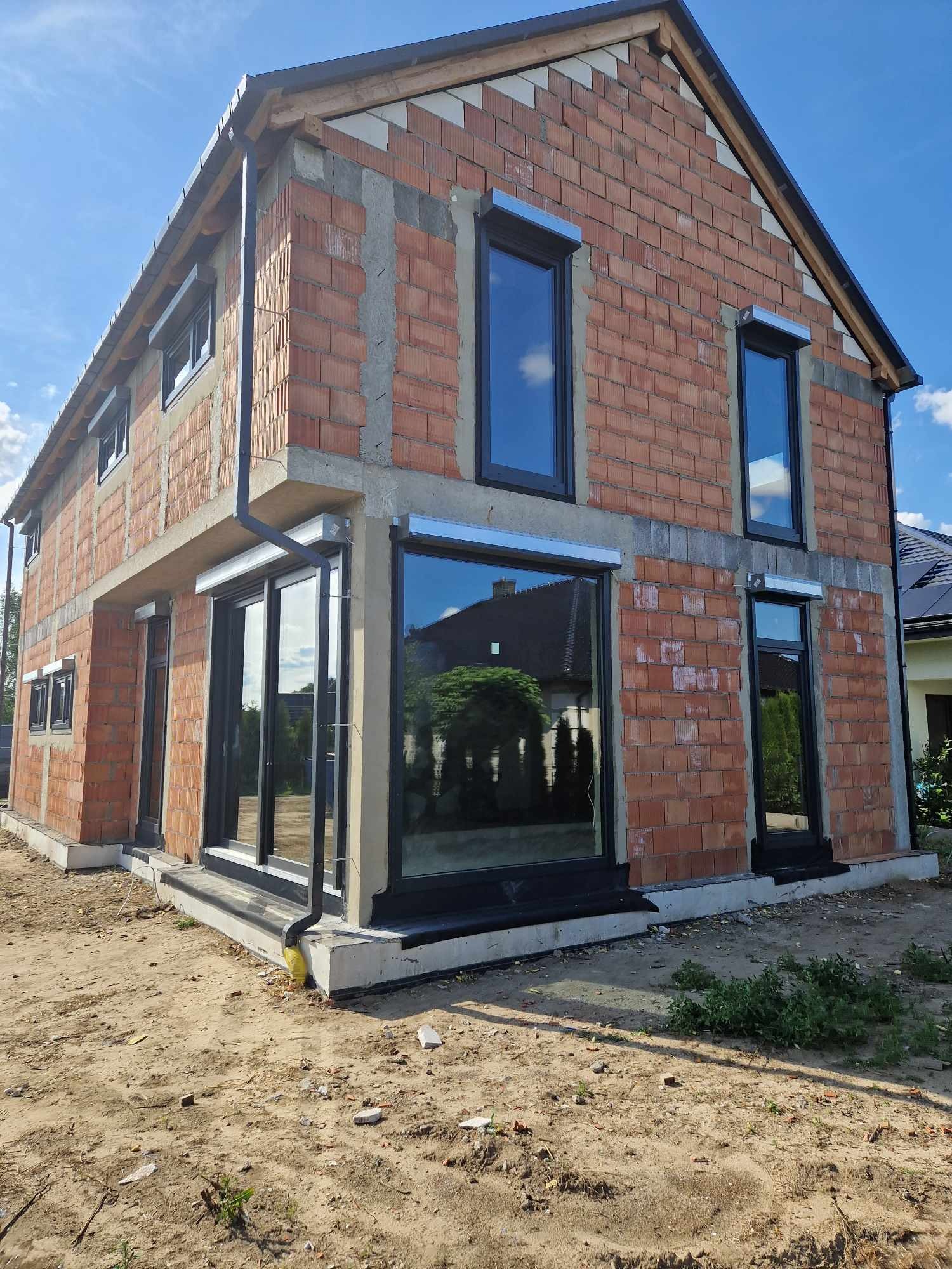 Surowy stan budynku jednorodzinnego z zamontowanymi czarnymi oknami i roletami zewnętrznymi, widok z zewnątrz na plac budowy z piaskiem.