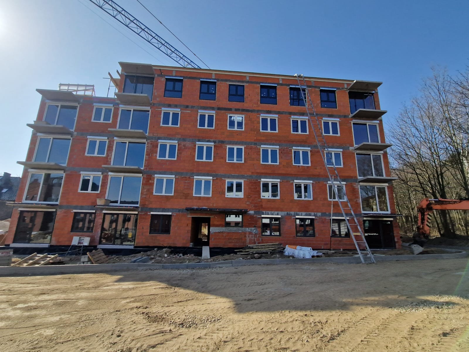 Budynek w trakcie budowy z zamontowanymi oknami o białych i ciemnych ramach, widoczna drabina oparta o ścianę, teren budowy z piaskiem i częściowo odsłoniętą cegłą.