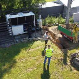 Widok z góry na instalację przemysłowej pompy ciepła w ogrodzie, z widocznym wykopem, minikoparką, traktorem z przyczepą i dwoma pracownikami w trakcie prac instalacyjnych.
