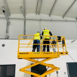 Dw&oacute;ch monter&oacute;w w ż&oacute;łtych kamizelkach, stojących na podnośniku Haulotte Compact 8, instaluje rury wentylacyjne pod sufitem w hali przemysłowej z białymi ścianami z bloczk&oacute;w i widocznymi elementami...