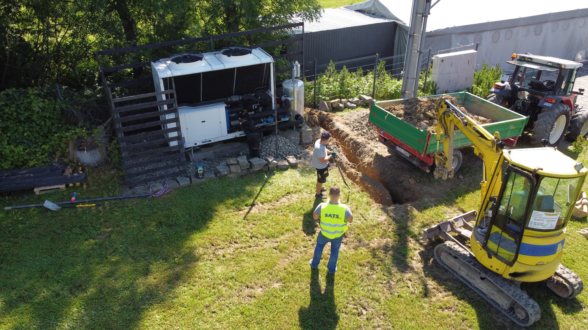 Widok z góry na instalację przemysłowej pompy ciepła w ogrodzie, z widocznym wykopem, minikoparką, traktorem z przyczepą i dwoma pracownikami w trakcie prac instalacyjnych.