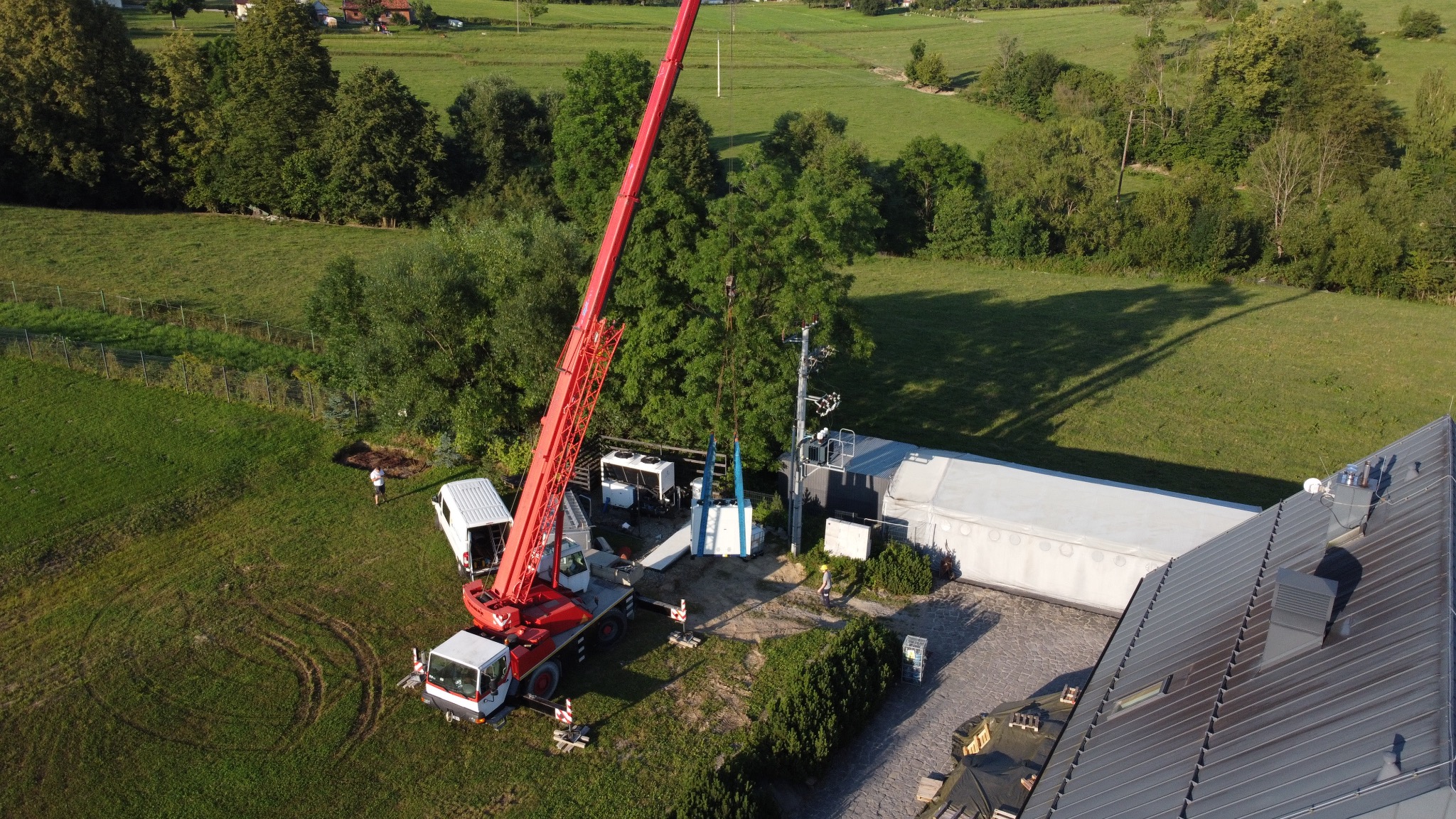Widok z drona: czerwony dźwig podnosi modułową jednostkę klimatyzacyjną na tle zielonego krajobrazu i budynku z szarym dachem.