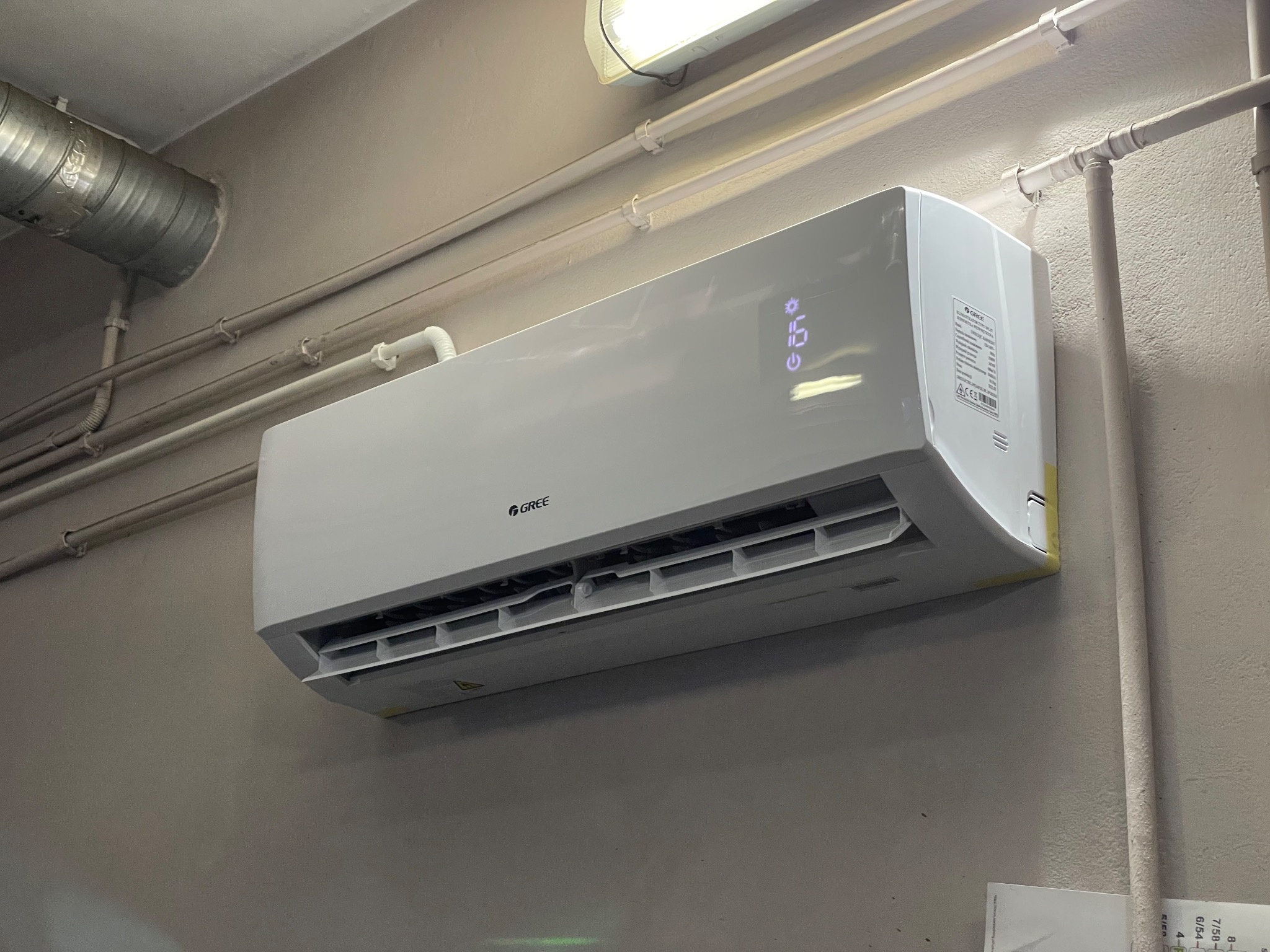 Biała jednostka wewnętrzna klimatyzatora marki Gree zamontowana na szarej ścianie, widoczne rury instalacyjne pod sufitem.