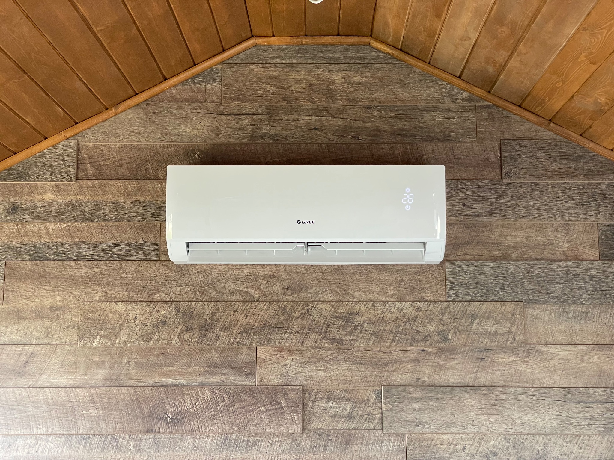 Biały klimatyzator marki Gree zamontowany na ścianie wyłożonej panelami imitującymi drewno, pod drewnianym sufitem w kształcie trójkąta.