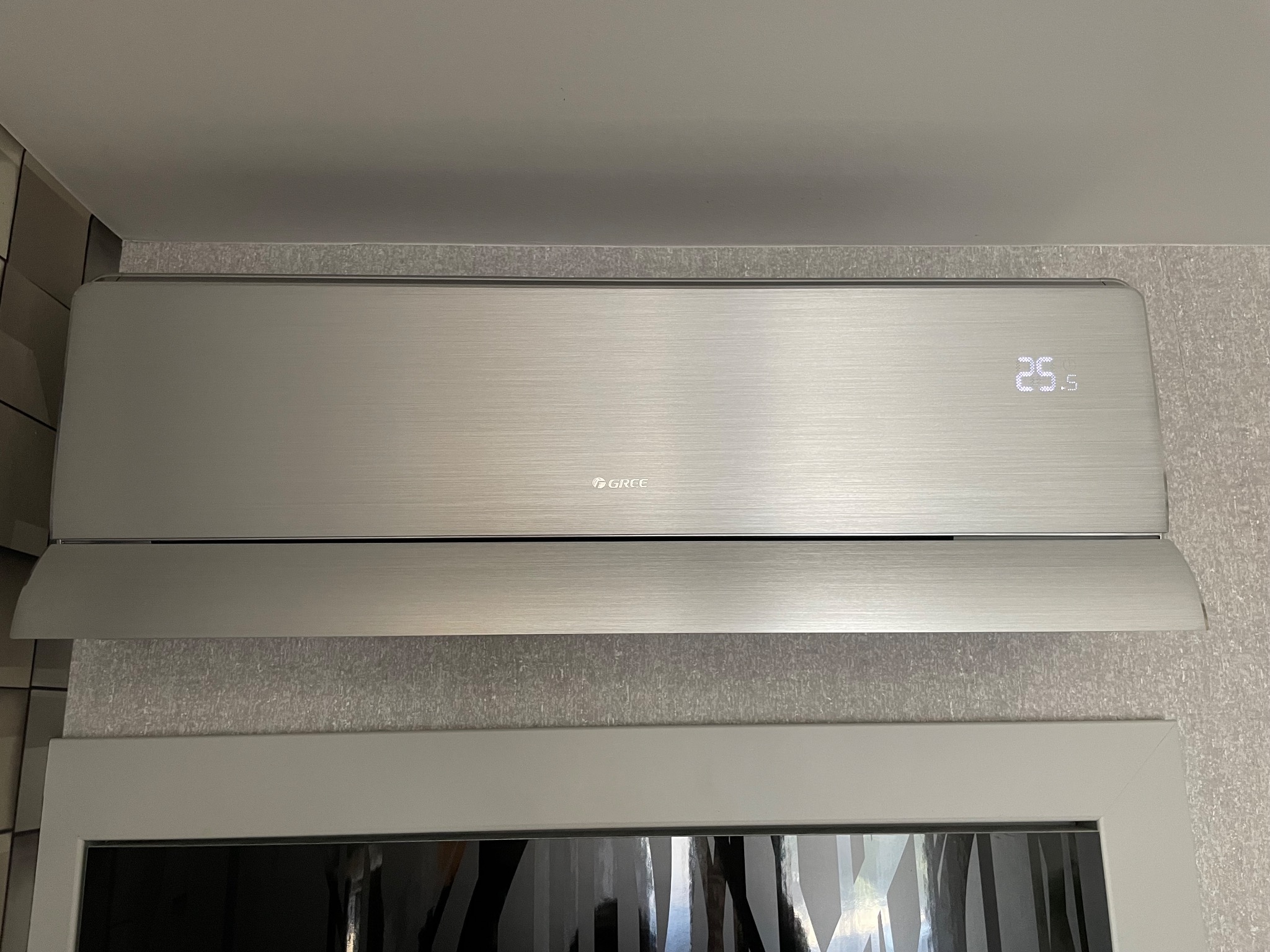 Szczegółowe ujęcie srebrnej klimatyzacji GREE zamontowanej na ścianie z tapetą, wyświetlającej temperaturę 25.5 stopni Celsjusza.