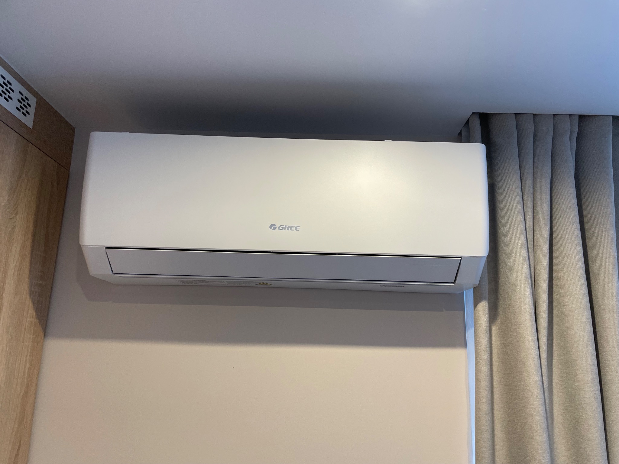 Biała jednostka wewnętrzna klimatyzacji marki Gree zamontowana na beżowej ścianie, obok zasłony w odcieniach szarości i elementu drewnianej zabudowy.