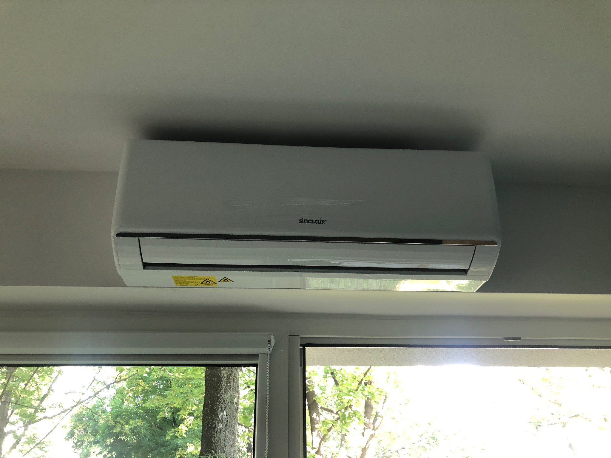 Biała klimatyzacja marki Sinclair zamontowana na suficie, widok z dołu, okno z widokiem na drzewa w tle.