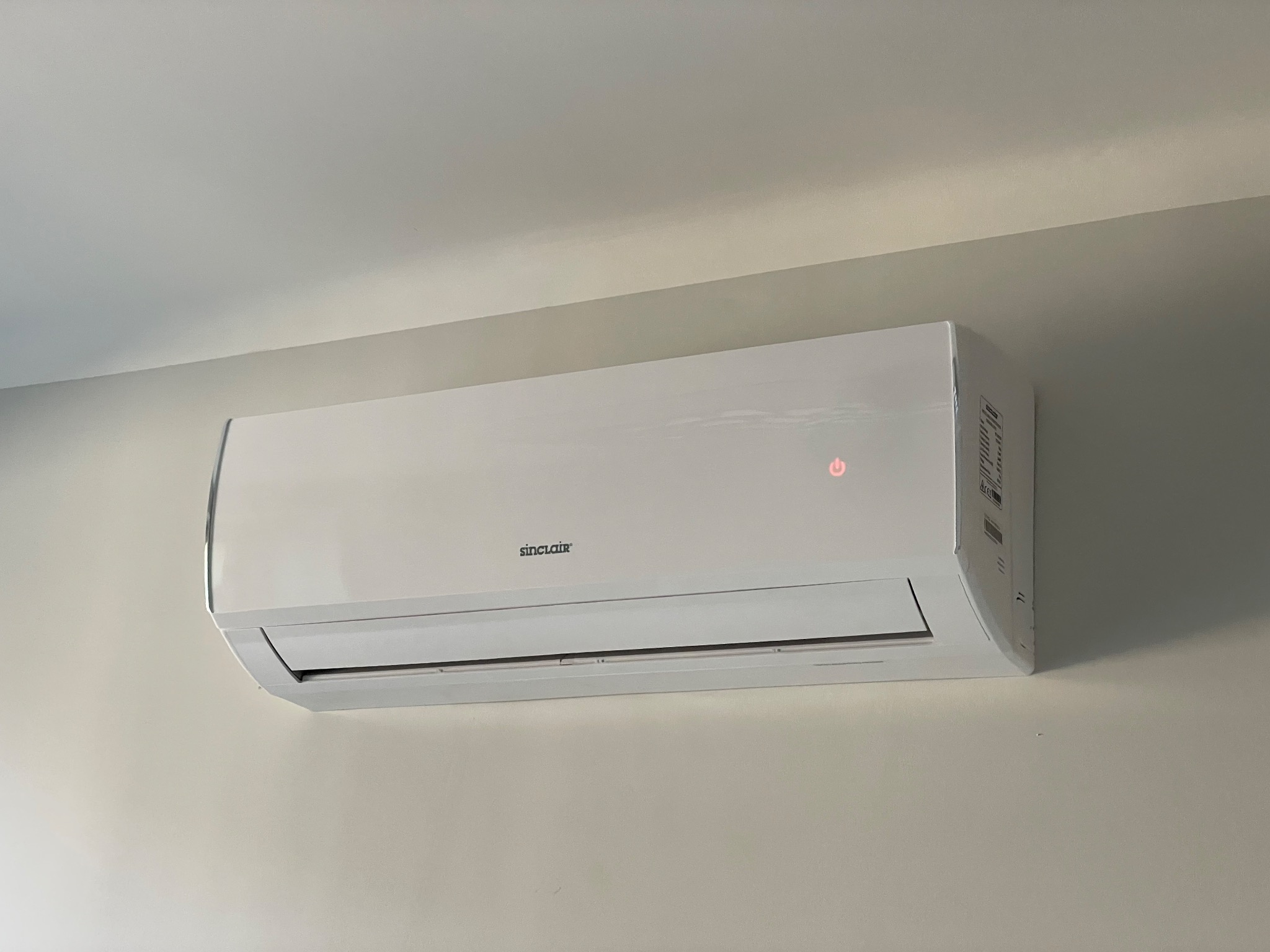 Biała klimatyzacja Sinclair zamontowana na jasnej ścianie, widoczna dioda sygnalizacyjna w prawym górnym rogu urządzenia.