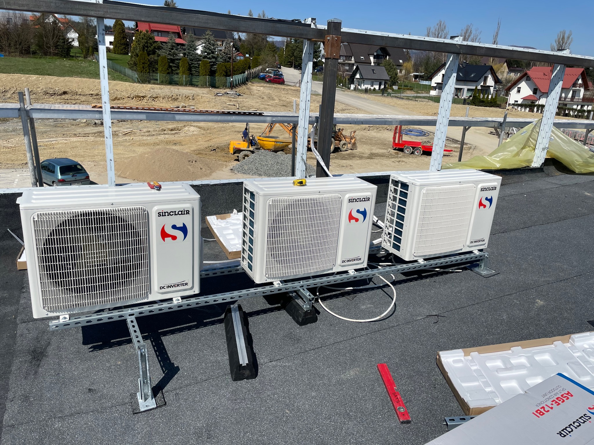 Trzy zewnętrzne jednostki klimatyzacji Sinclair DC Inverter zamontowane na metalowej konstrukcji na dachu, z widokiem na plac budowy z koparką i domami w tle.