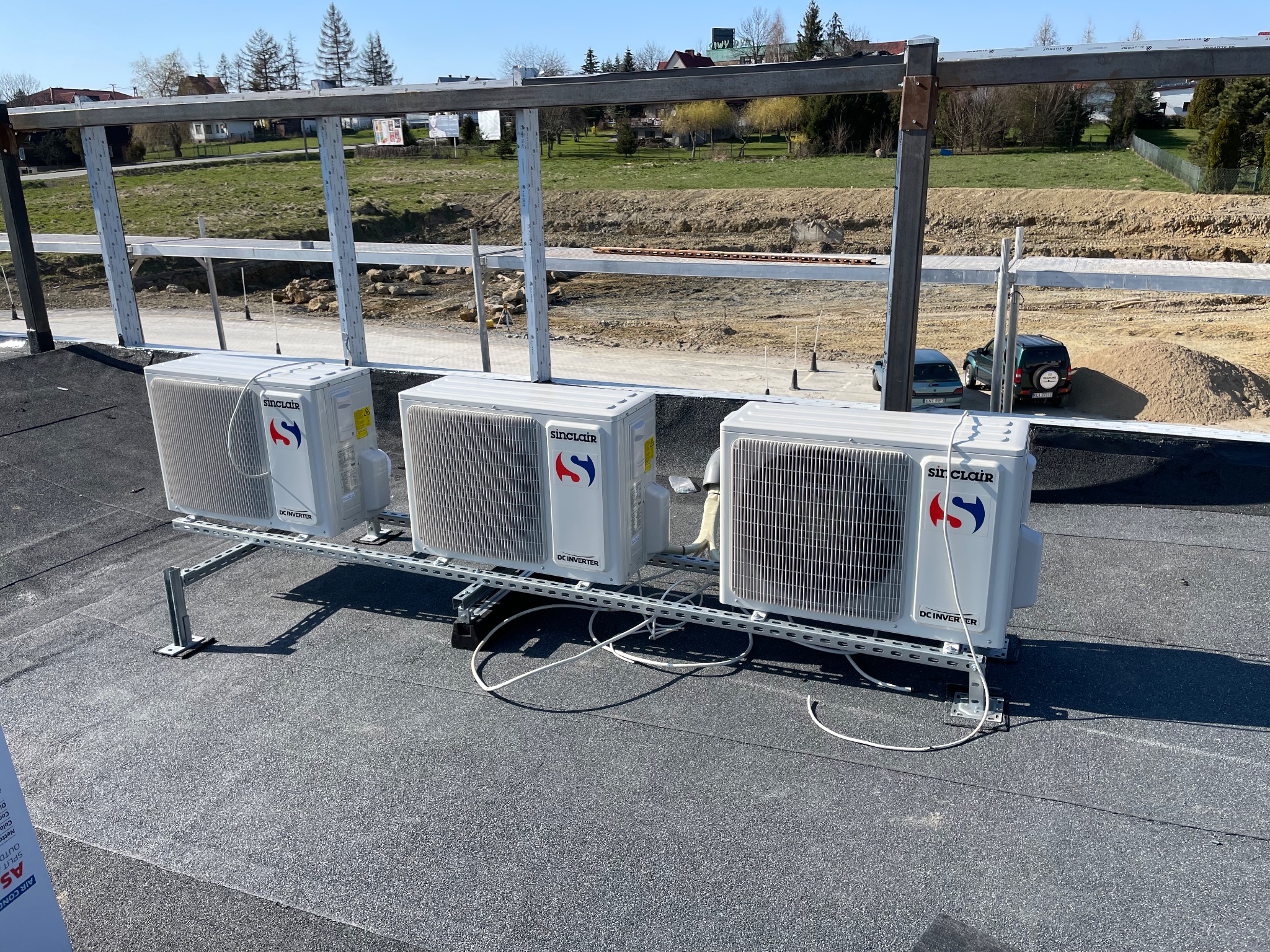 Trzy jednostki zewnętrzne klimatyzacji Sinclair DC Inverter zamontowane na dachu budynku w trakcie instalacji, widoczne elementy konstrukcji wsporczej i teren budowy w tle.
