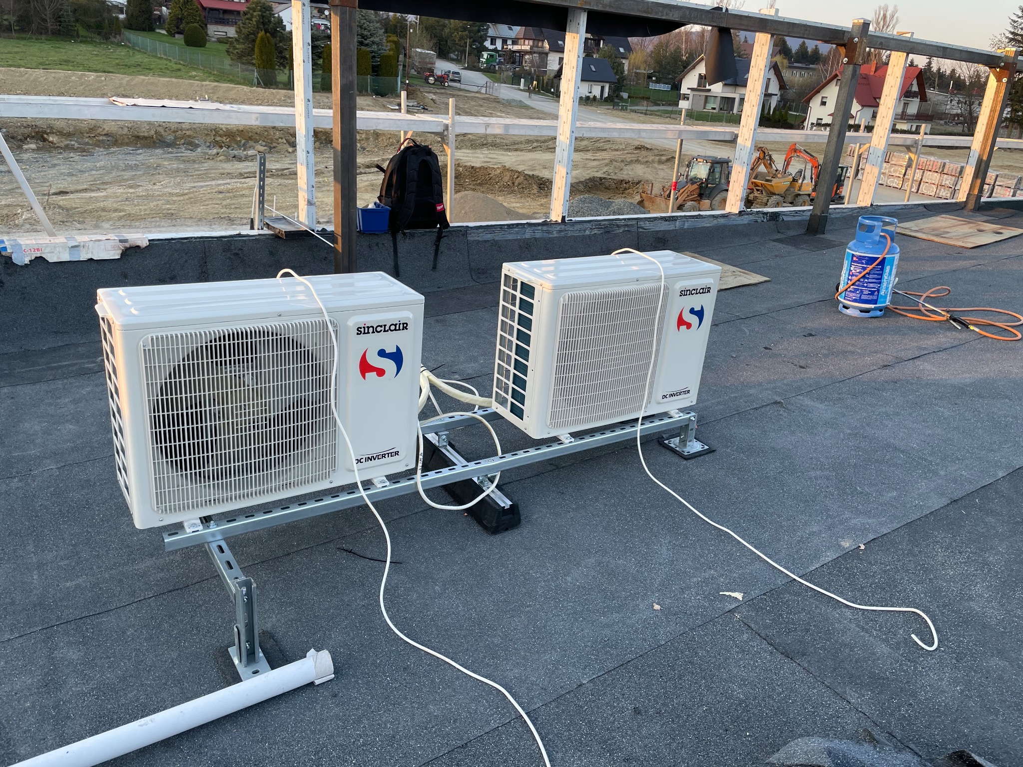 Dwie jednostki zewnętrzne klimatyzacji Sinclair zamontowane na dachu budynku w trakcie budowy, widoczna butla gazowa i sprzęt budowlany w tle.
