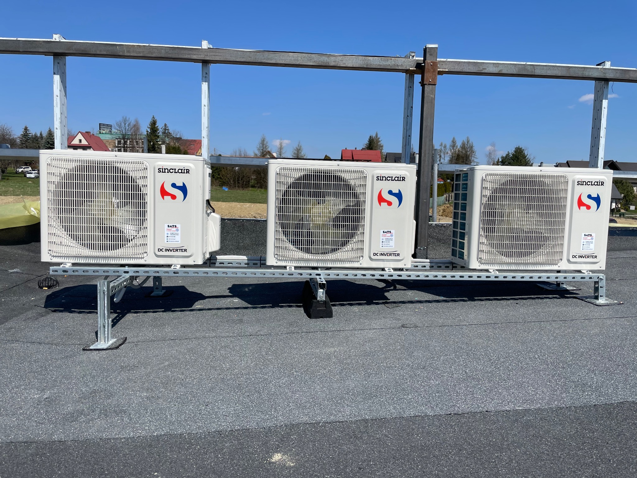 Trzy zewnętrzne jednostki klimatyzacyjne Sinclair DC Inverter, zamontowane na metalowej konstrukcji na płaskim dachu, widoczne budynki w tle pod błękitnym niebem.
