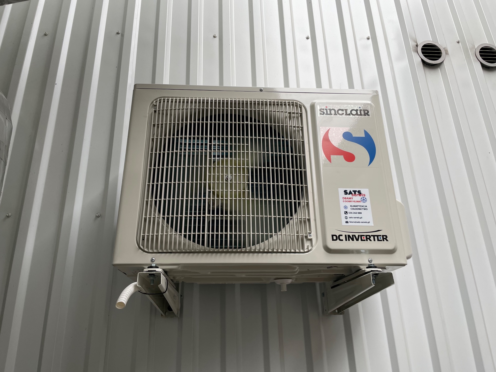 Zamontowany klimatyzator Sinclair DC Inverter na białej, metalowej ścianie z widocznymi śrubami i elementami instalacji.