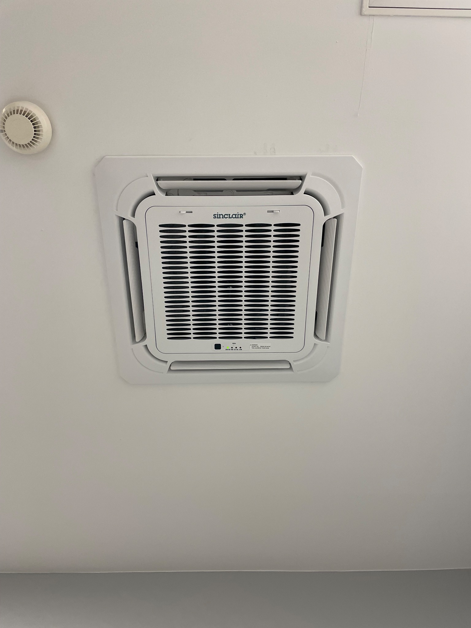 Kasetonowa jednostka klimatyzacji Sinclair zamontowana w suficie, widoczne diody sygnalizacyjne i kratka wentylacyjna, obok czujnik dymu.