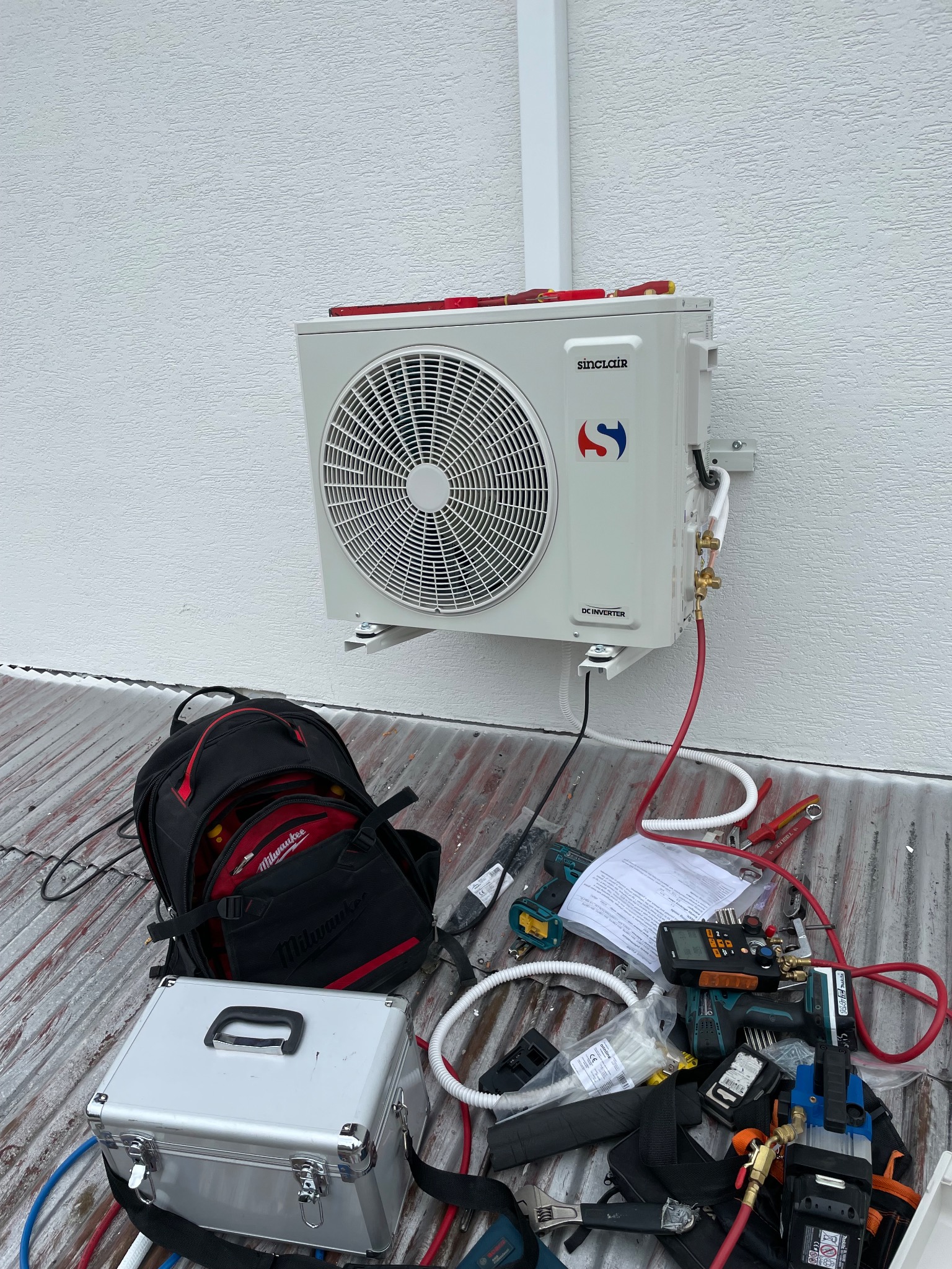 Zamontowana zewnętrzna jednostka klimatyzacji Sinclair na białej elewacji budynku, widoczne narzędzia instalacyjne, torba Milwaukee oraz srebrna walizka narzędziowa na dachu.