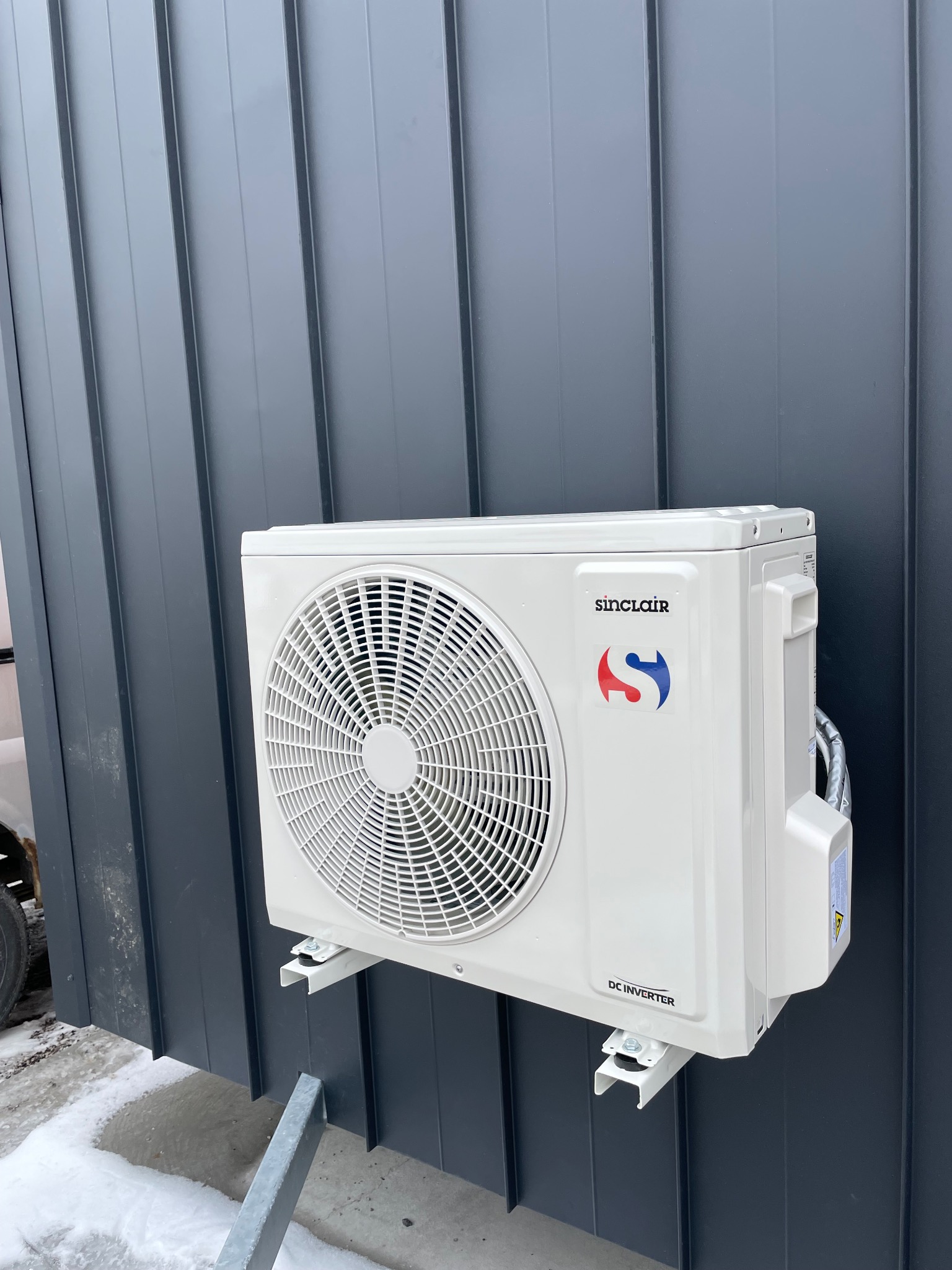 Biała jednostka zewnętrzna klimatyzacji Sinclair zamontowana na szarej, metalowej elewacji budynku. Widoczny śnieg na ziemi.