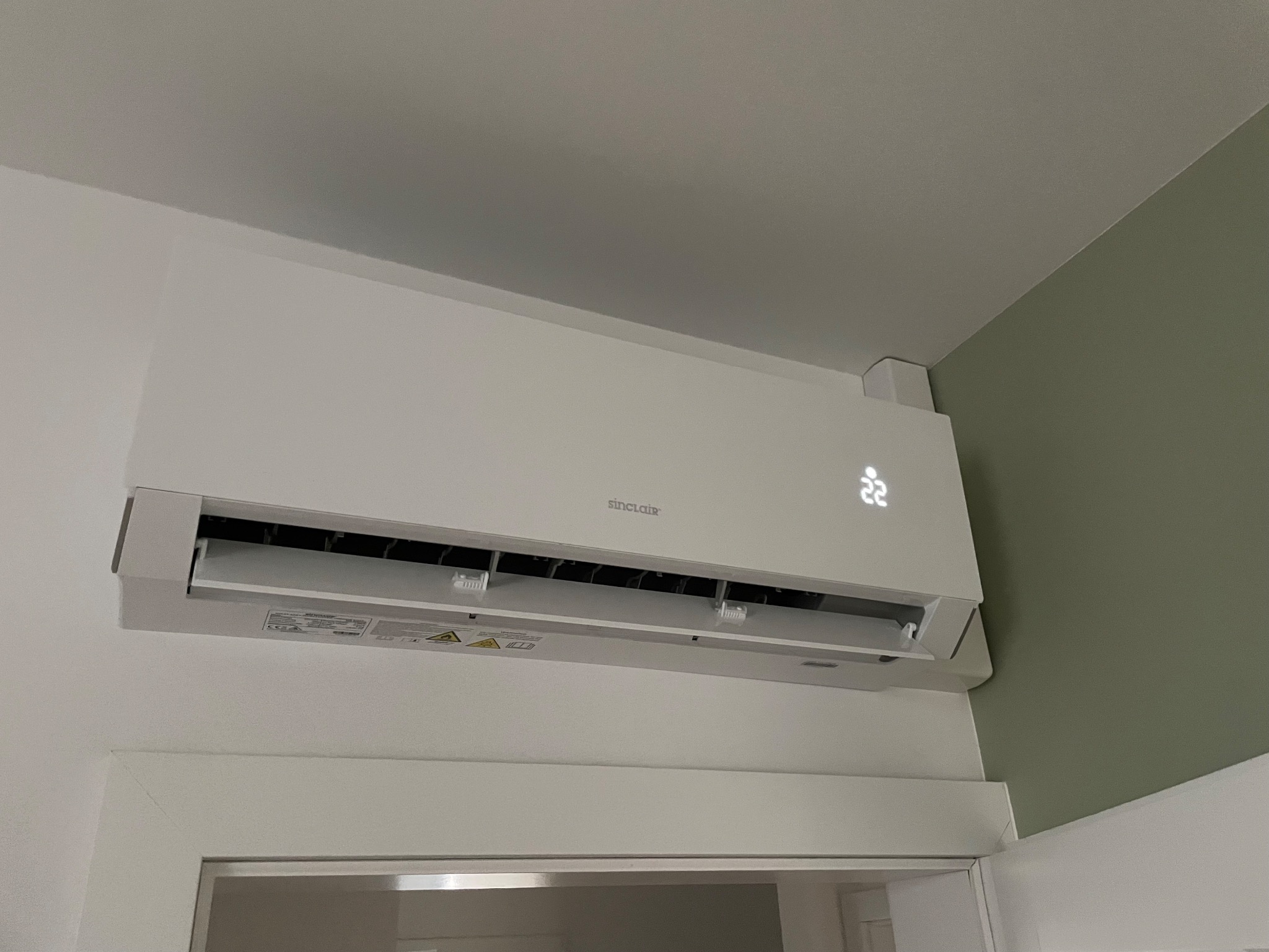 Biały klimatyzator Sinclair zamontowany na ścianie nad drzwiami, z wyświetlaczem LED wskazującym temperaturę 22 stopnie Celsjusza, na tle białej i oliwkowozielonej ściany.