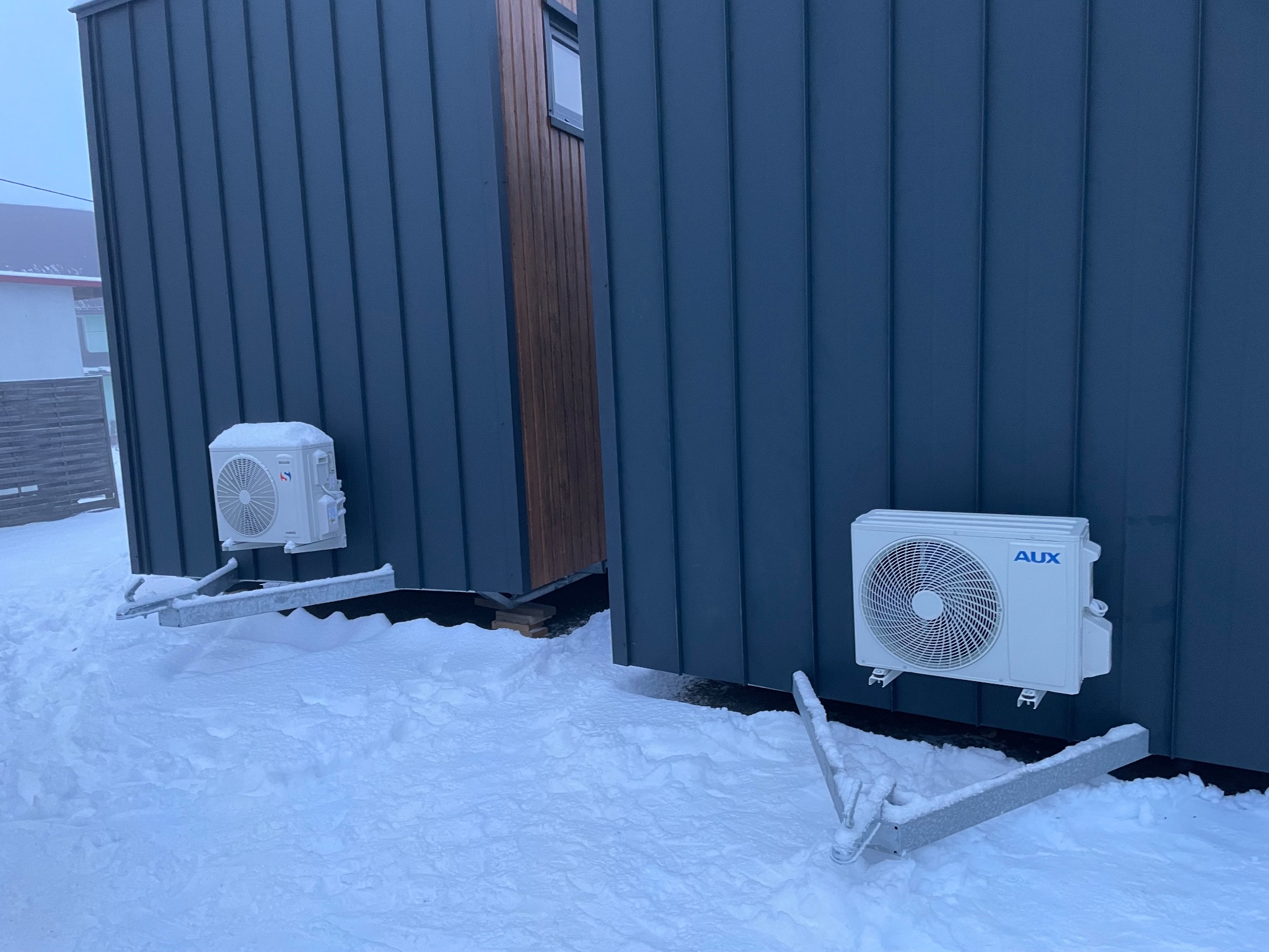 Dwa modułowe domki z zamontowanymi klimatyzatorami AUX na zewnątrz w zimowej scenerii, pokryte śniegiem, widoczne metalowe konstrukcje wsporcze.