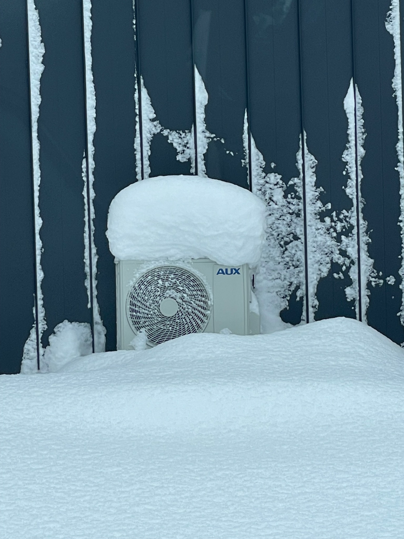 Agregat klimatyzacji marki AUX zasypany śniegiem, widoczny na tle ciemnej, pionowo ułożonej elewacji pokrytej śnieżnym nalotem.
