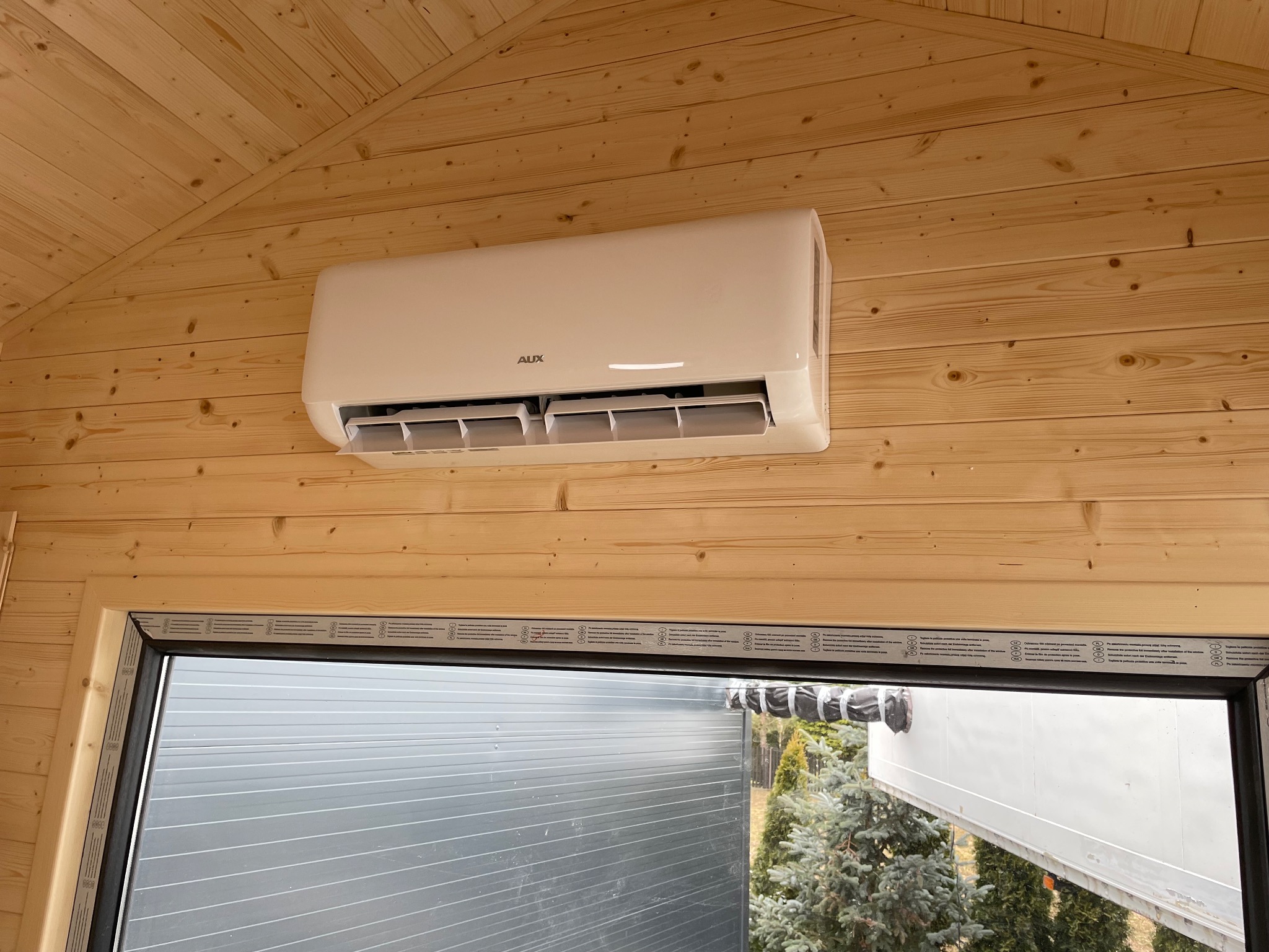 Klimatyzator marki AUX zamontowany w pomieszczeniu wykończonym boazerią w Rabce-Zdroju, widok przez okno na zewnątrz.