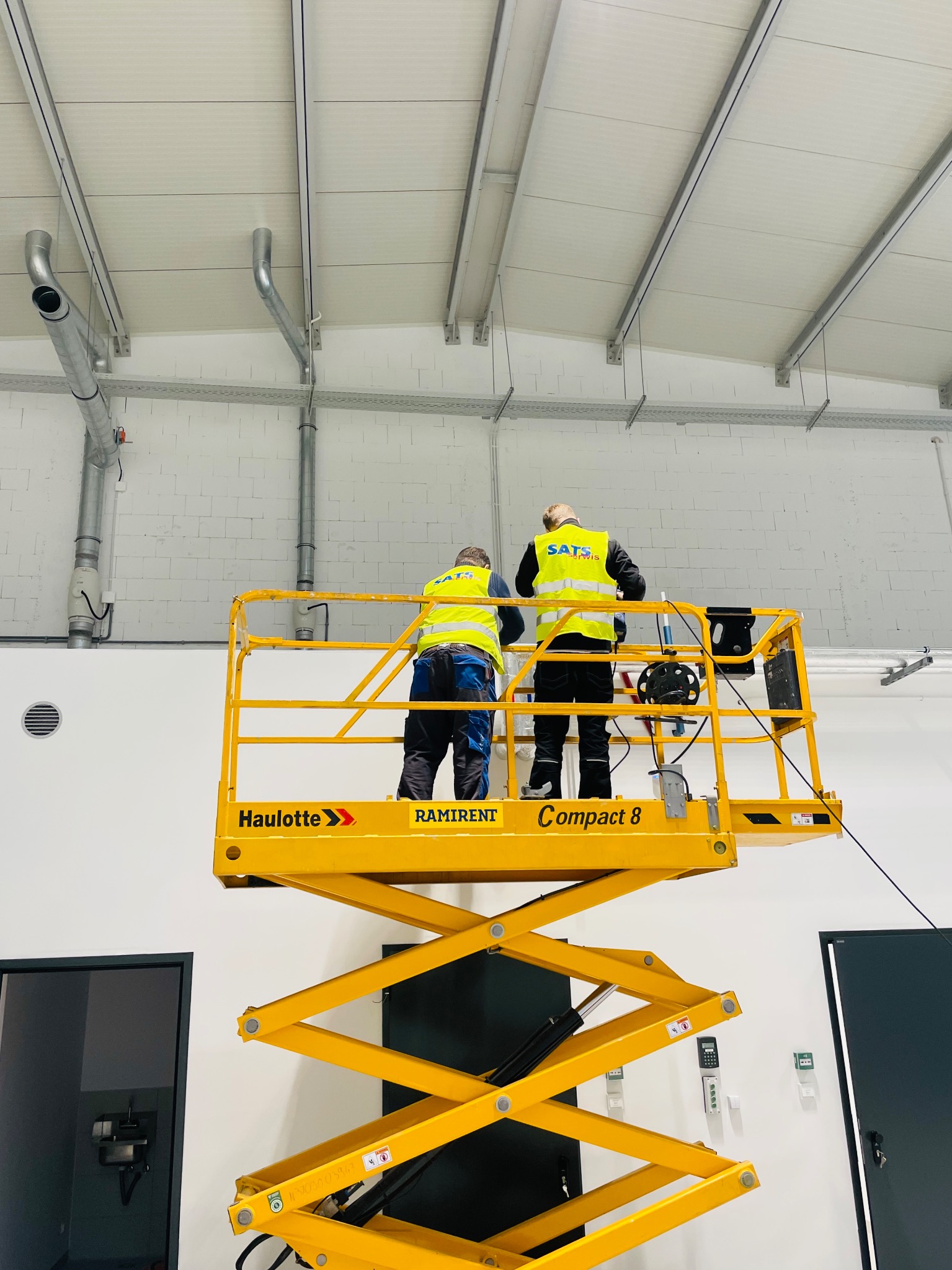 Dwóch monterów w żółtych kamizelkach, stojących na podnośniku Haulotte Compact 8, instaluje rury wentylacyjne pod sufitem w hali przemysłowej z białymi ścianami z bloczków i widocznymi elementami...