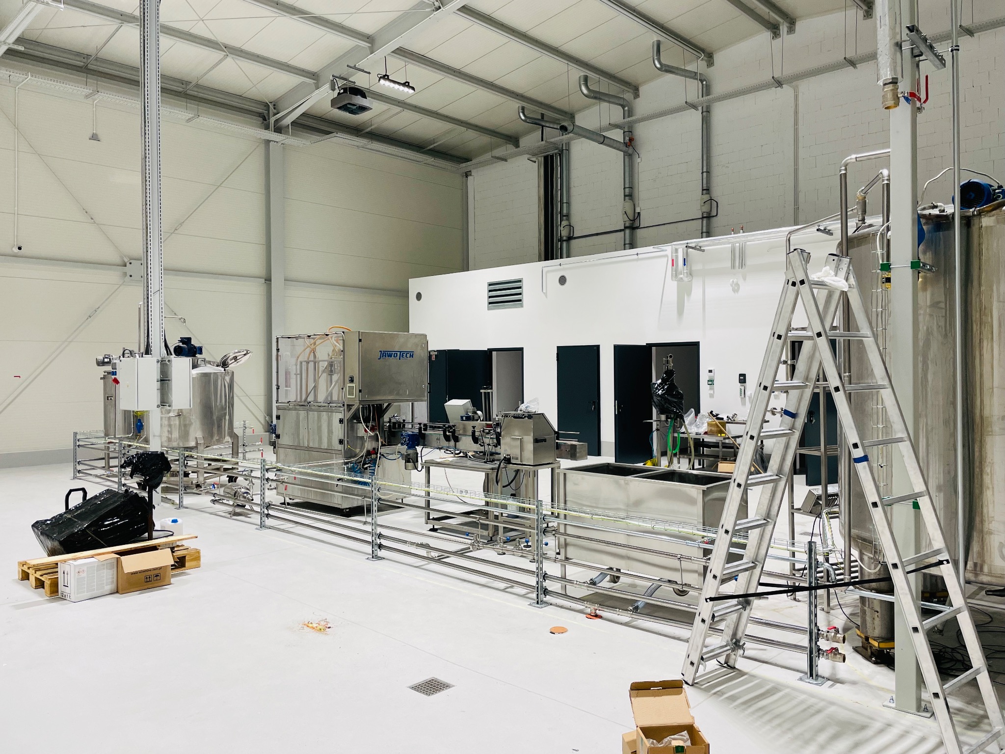 Wnętrze hali produkcyjnej z zainstalowaną linią technologiczną, widoczne rury, zbiorniki ze stali nierdzewnej, drabina aluminiowa i elementy montażowe, w tle trzy pary drzwi.