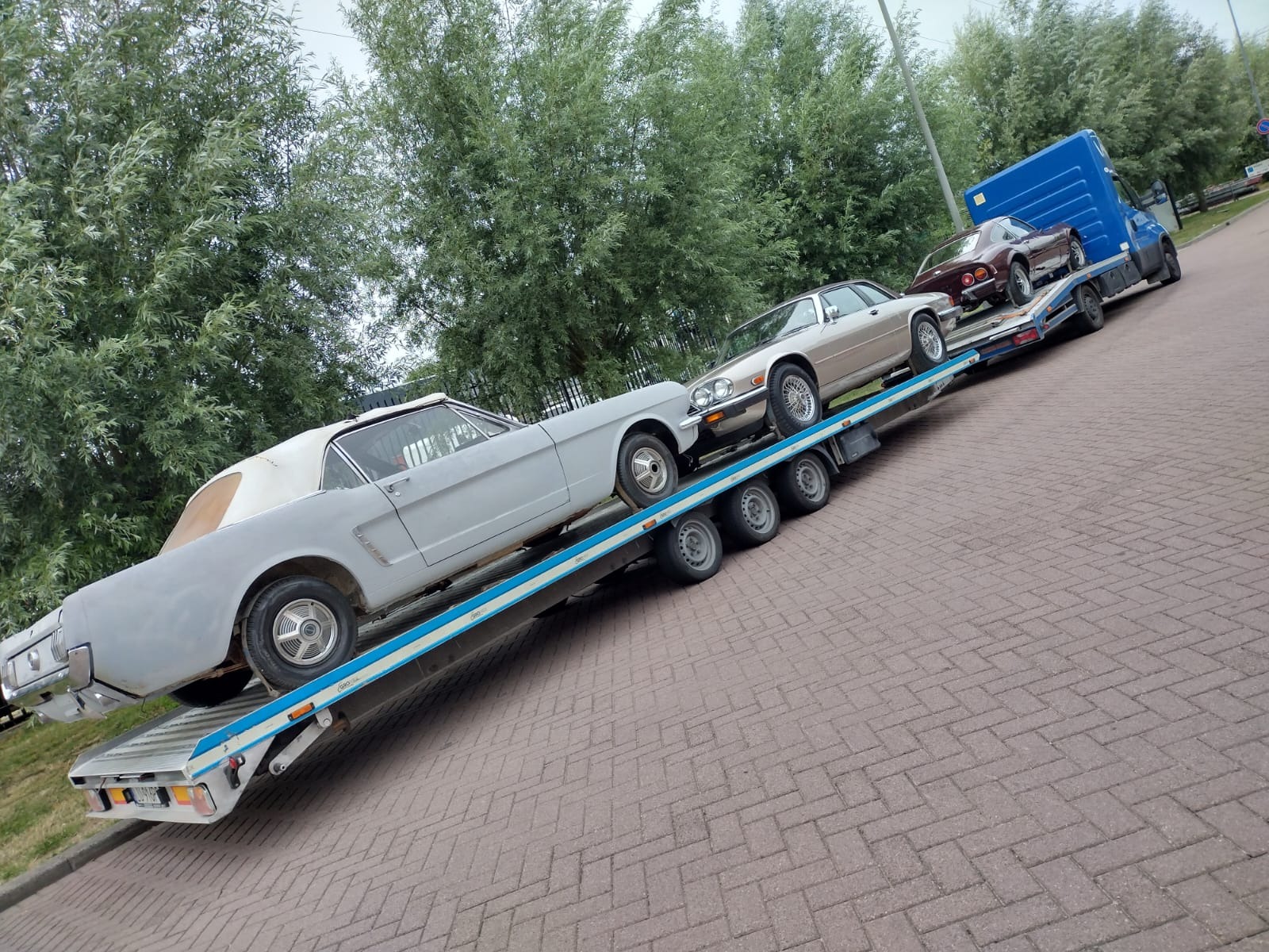 Laweta przewozi trzy zabytkowe samochody: jasnoszarego Forda Mustanga, beżowego Jaguara XJ i bordowego samochodu sportowego.