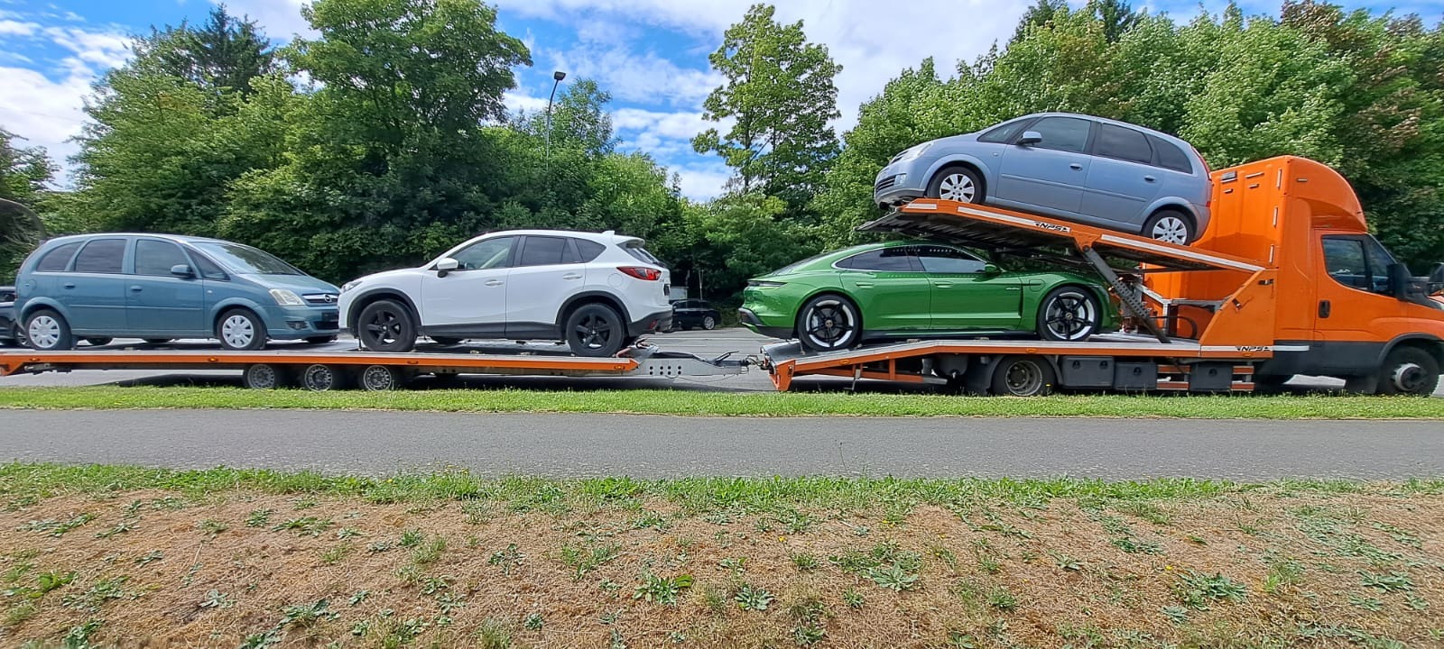 Pomarańczowa laweta z dwoma poziomami transportuje cztery samochody: niebieski minivan, biały SUV, zielony sportowy samochód i ponownie niebieski minivan na górnym poziomie.