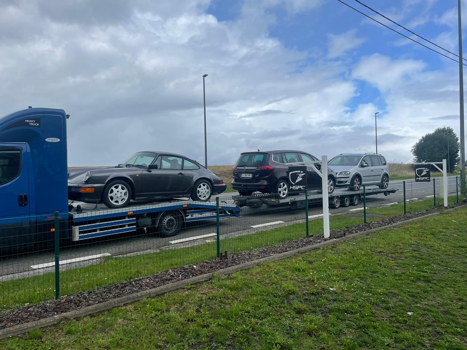 Laweta przewozi trzy samochody: szare Porsche 911, czarny Opel Zafira i srebrny Volkswagen Touran, wszystkie z logo HSPC na boku.