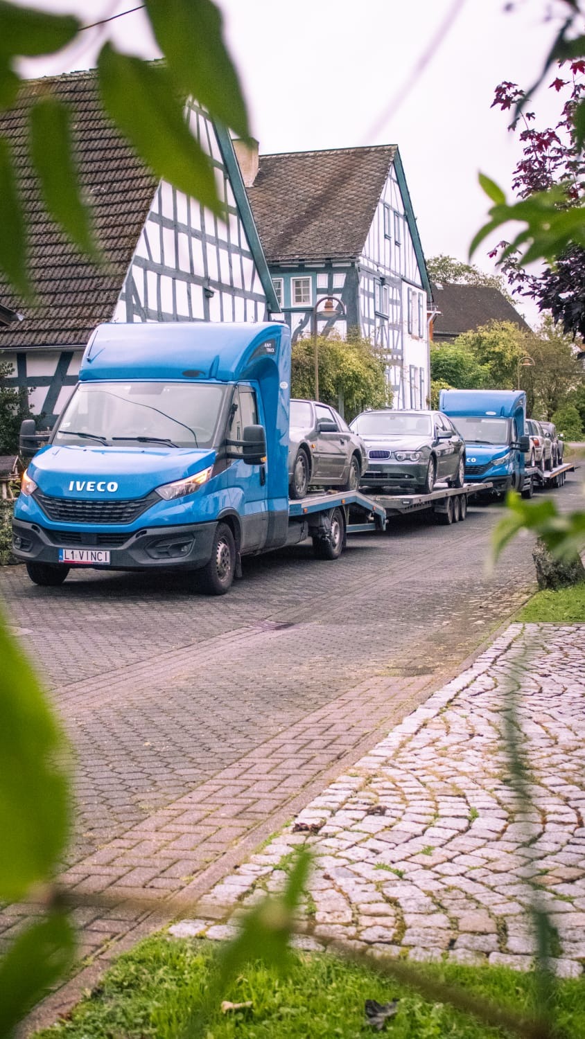 Dwa niebieskie samochody ciężarowe z naczepami przewożące samochody osobowe na tle domów z muru pruskiego, widok z perspektywy ulicy wyłożonej kostką.
