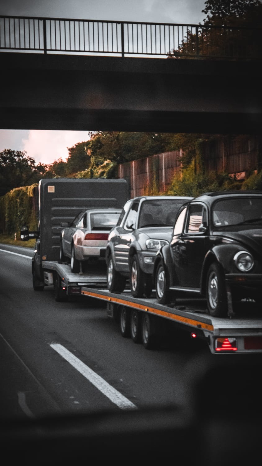 Laweta przewozi trzy samochody: srebrne sportowe auto, szarego SUV-a i czarnego Volkswagena Garbusa pod wiaduktem.