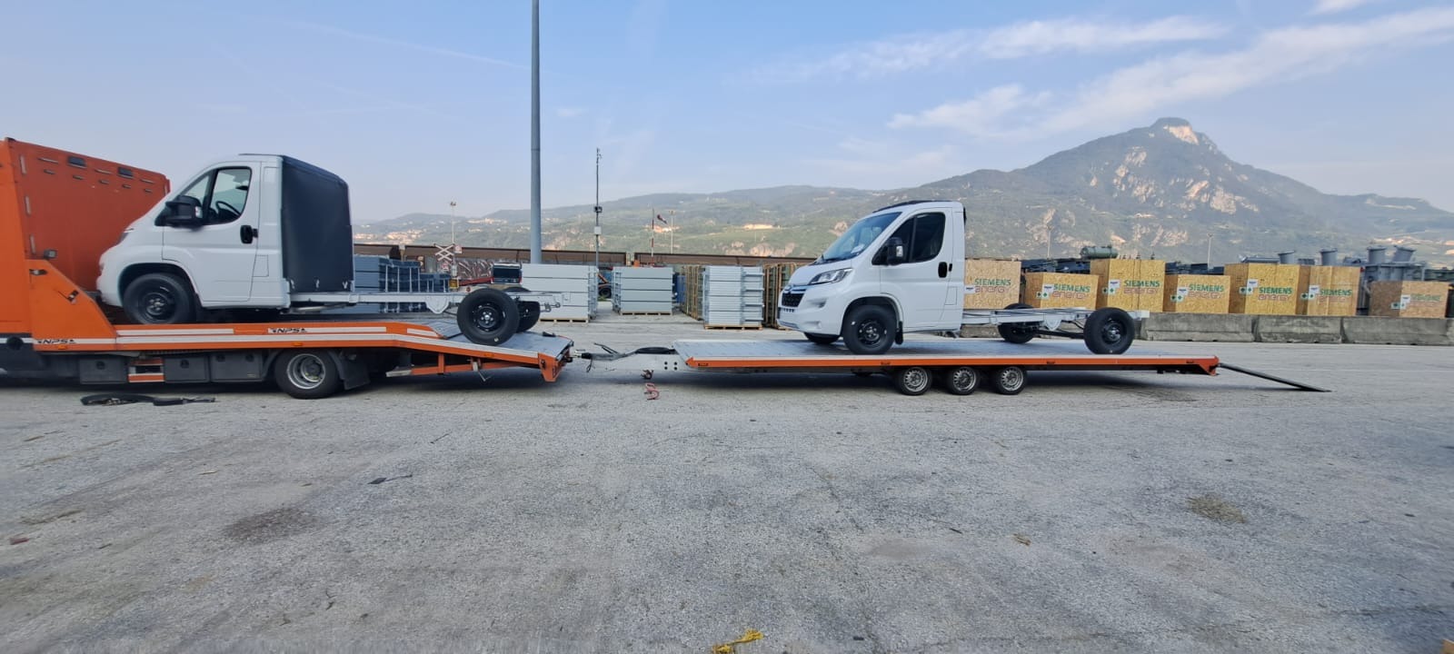 Pomarańczowa laweta z białym samochodem dostawczym na platformie, drugi biały samochód dostawczy na platformie holowniczej, w tle góry.