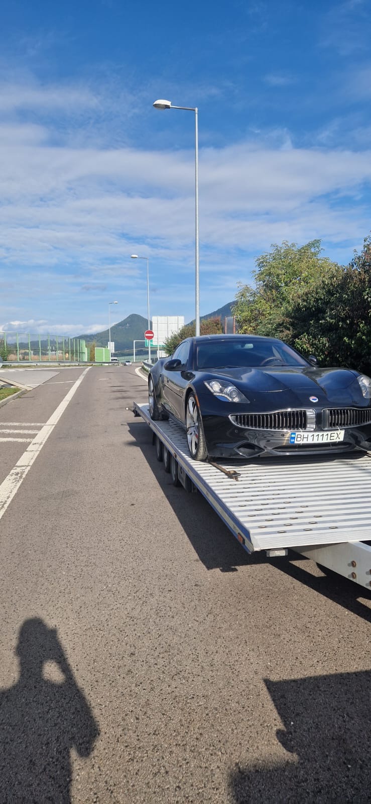 Czarny samochód marki Fisker z widoczną tablicą rejestracyjną BH 1111 EX, przewożony na niskopodwoziowej platformie transportowej na tle krajobrazu z zielenią i błękitnym niebem z chmurami.