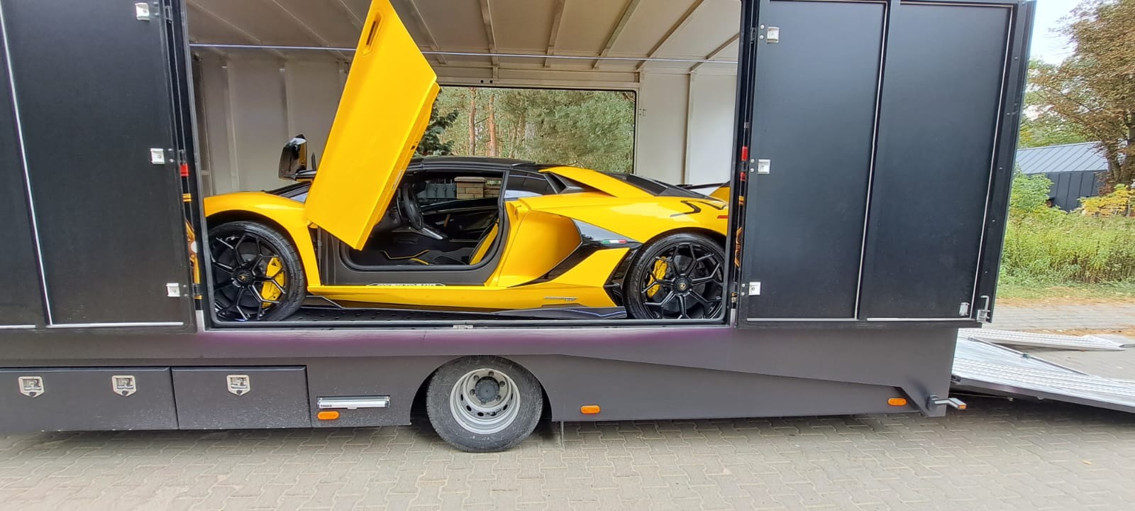 Żółty Lamborghini Aventador z otwartymi drzwiami, umieszczony wewnątrz czarnej, specjalistycznej przyczepy transportowej z opuszczoną rampą, na tle zieleni.