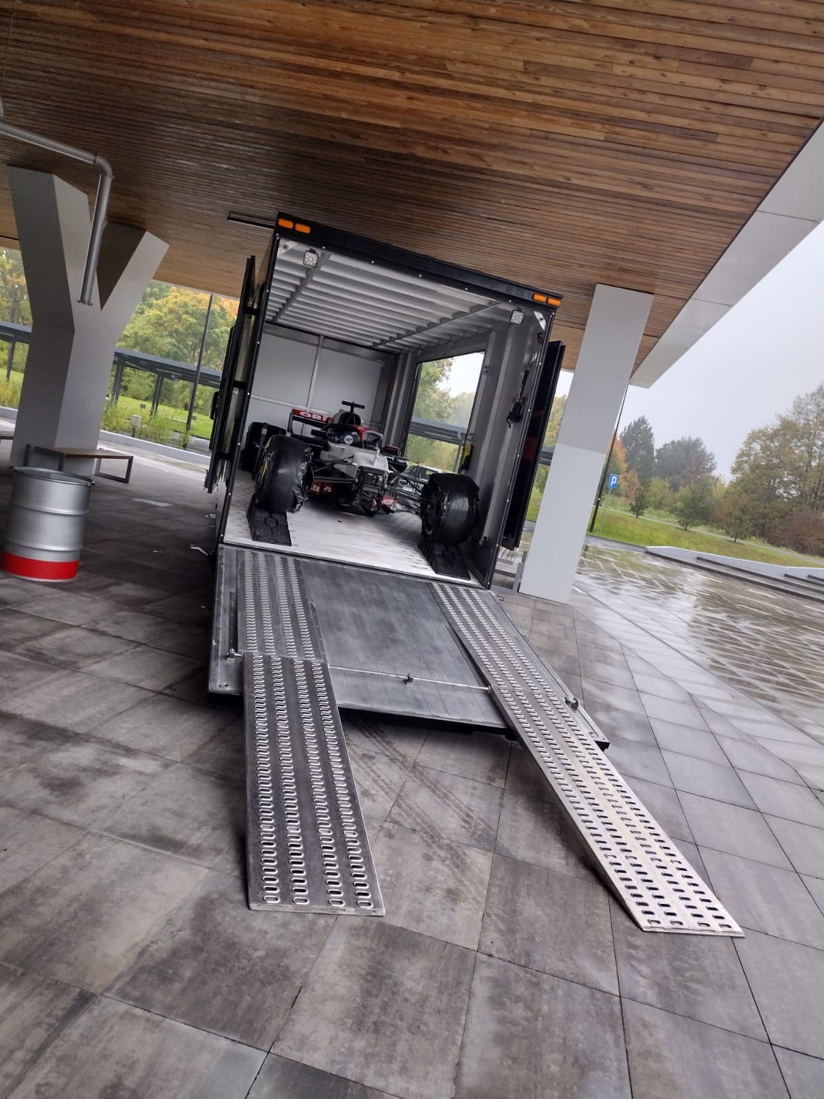 Samochód wyścigowy Formuły 1 umieszczony wewnątrz naczepy samochodu ciężarowego z rozłożonymi rampami, na zewnątrz nowoczesny budynek z drewnianym sufitem i betonowymi kolumnami, pochmurny dzień.