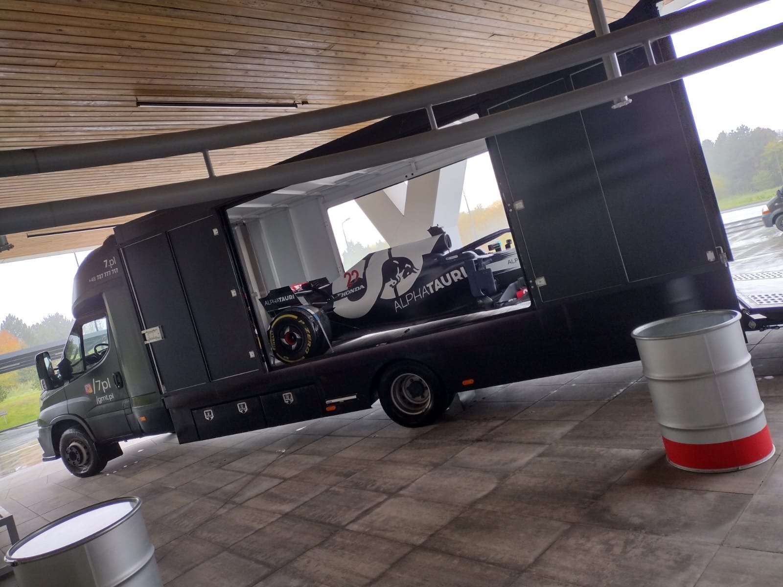 Czarny transportowy samochód ciężarowy z logiem firmy 7.pl przewozi bolid Formuły 1 AlphaTauri z numerem 22, widoczny przez otwarte drzwi naczepy.