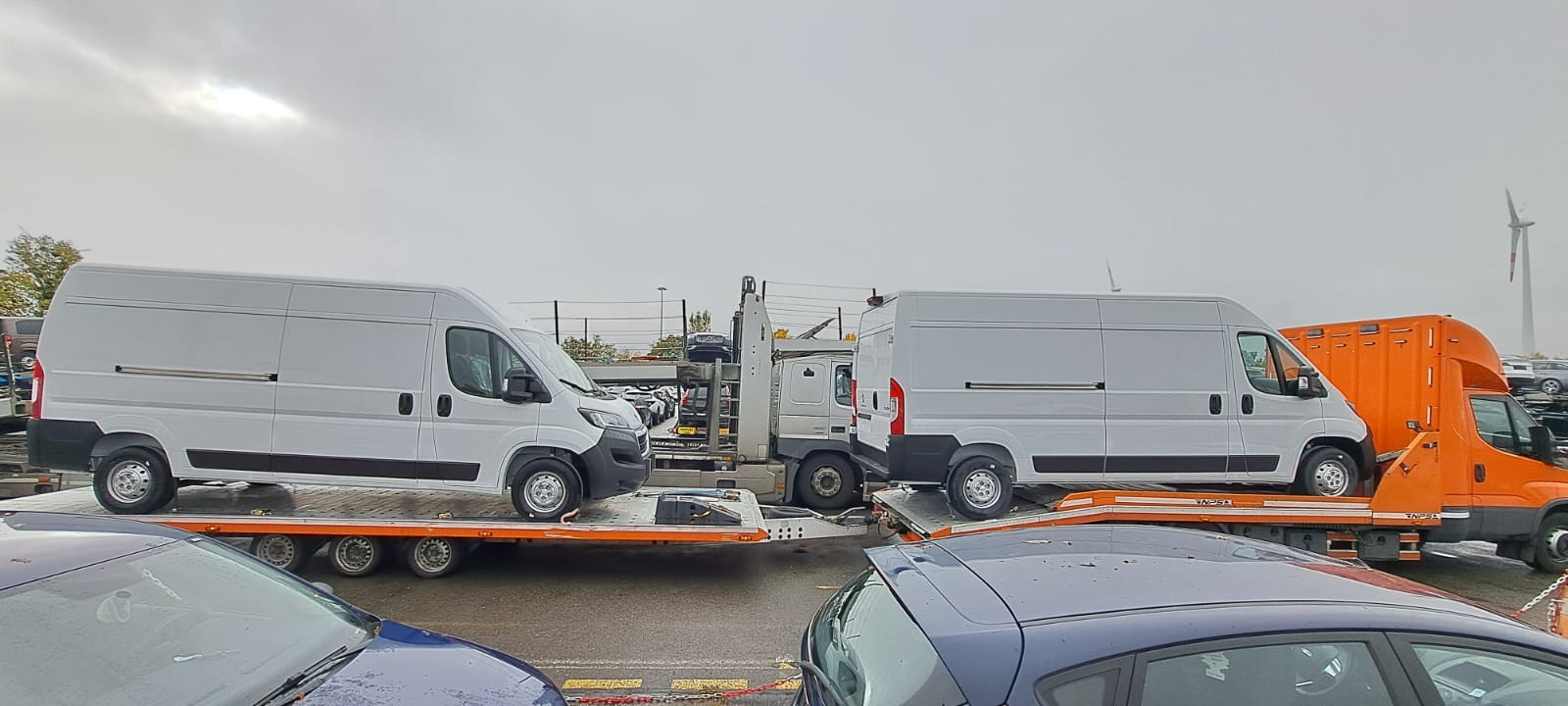 Dwa białe busy na pomarańczowej lawecie z widoczną turbiną w tle, transportowane w pochmurny dzień.