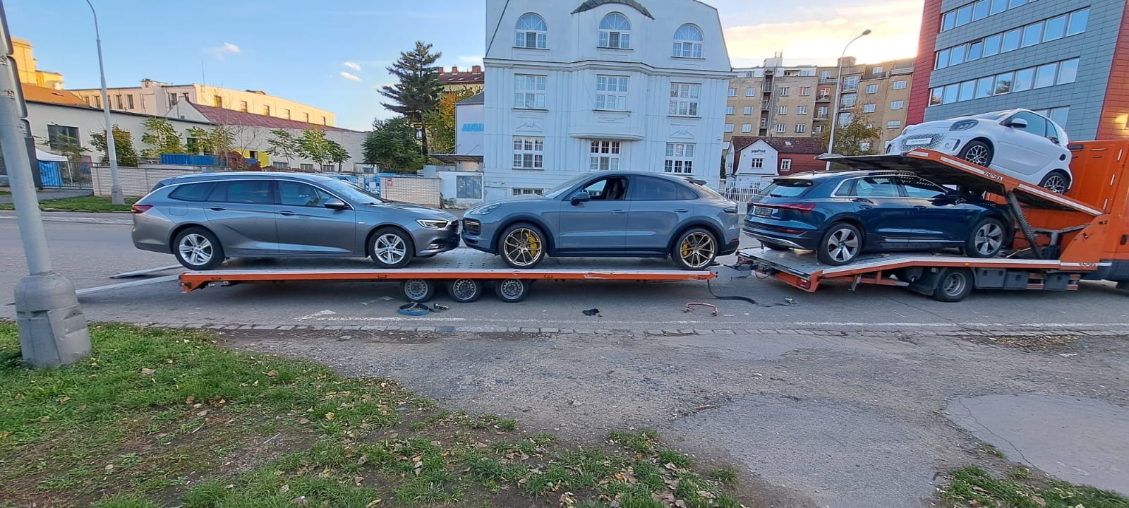 Pomarańczowa laweta z trzema samochodami: szary Opel Insignia kombi, szary Porsche Cayenne Coupe z żółtymi zaciskami hamulcowymi, niebieski Audi e-tron i biały Smart ForTwo na dwóch poziomach...