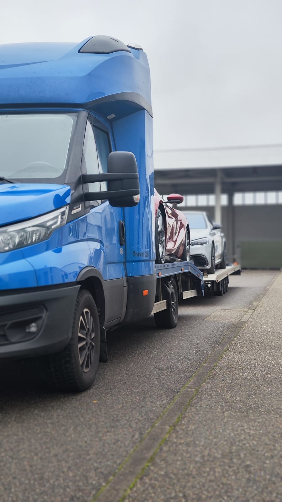 Niebieska laweta z dwoma sportowymi samochodami na platformie, czerwonym i białym, na szarym tle budynku przemysłowego.