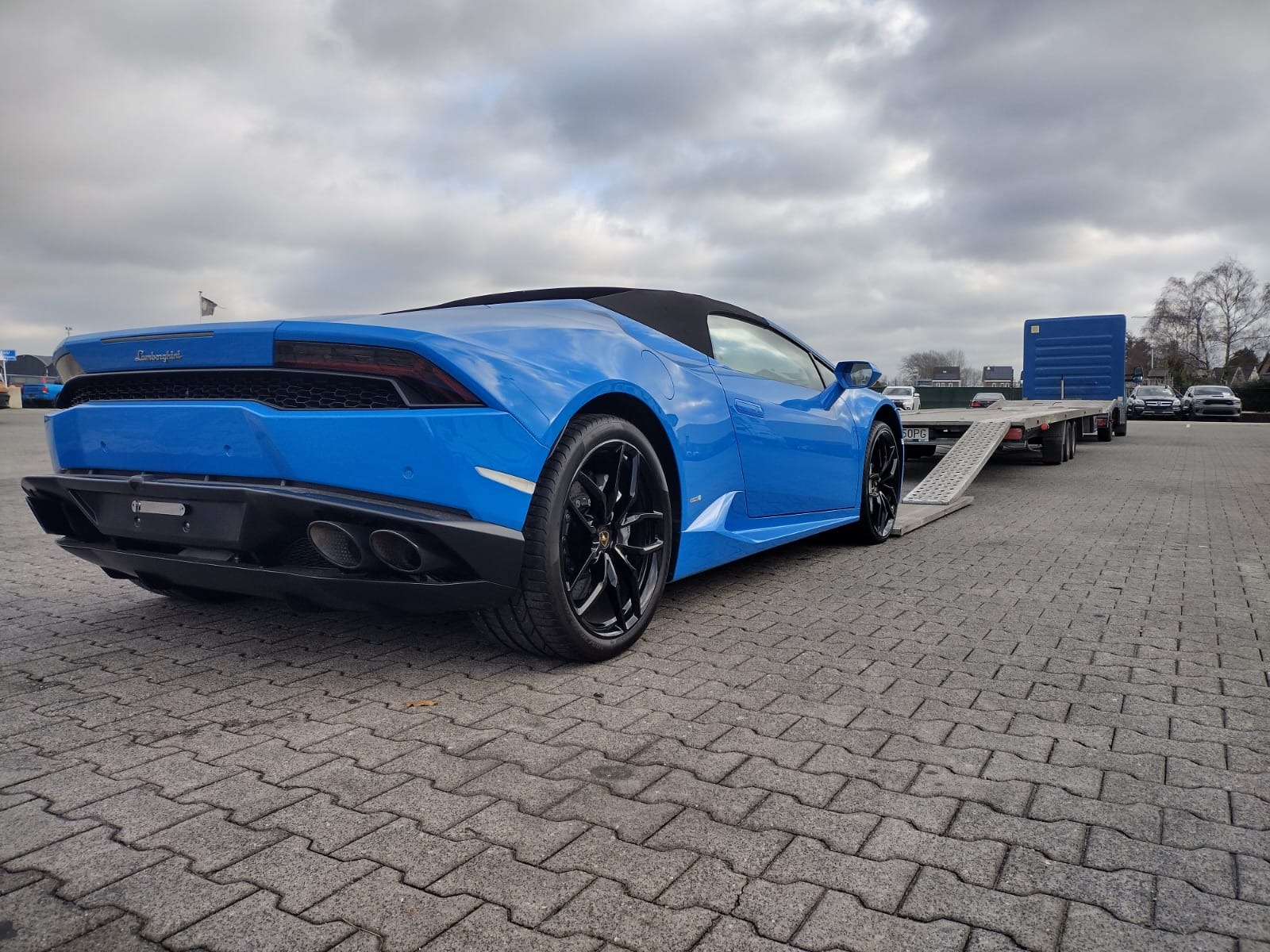 Błękitny Lamborghini Huracan Spyder na niskopodwoziowej lawecie z rampą, widok z tyłu, pochmurne niebo.